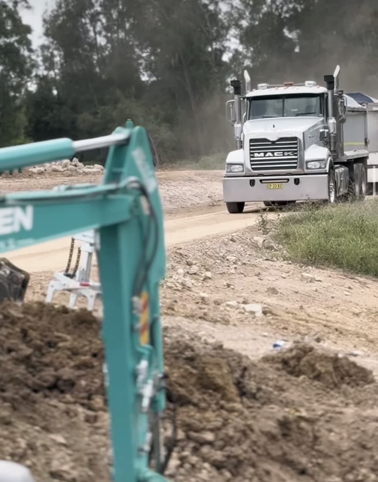 Teal Kobelco 5.5t excavator loading spoil into a white Mack tipper truck, dust rising