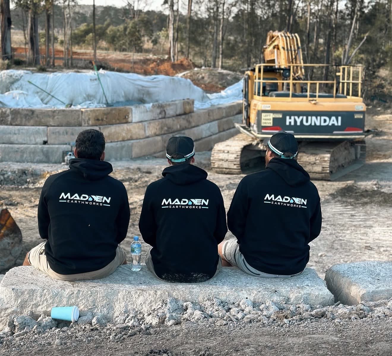 Three Madden Earthworks crew members in branded hoodies on a bulk earthworks site with a Hyundai 30t excavator behind